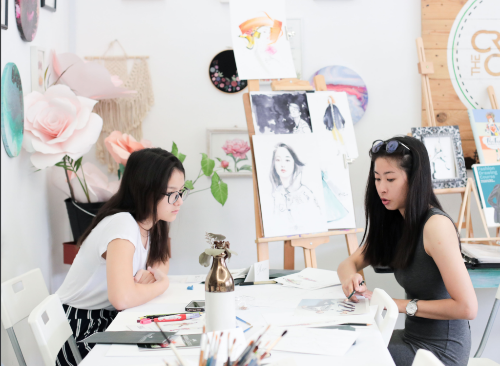 Two illustrators sketching fashion figures at a worktable, surrounded by framed watercolour and pencil artwork on the studio wall.